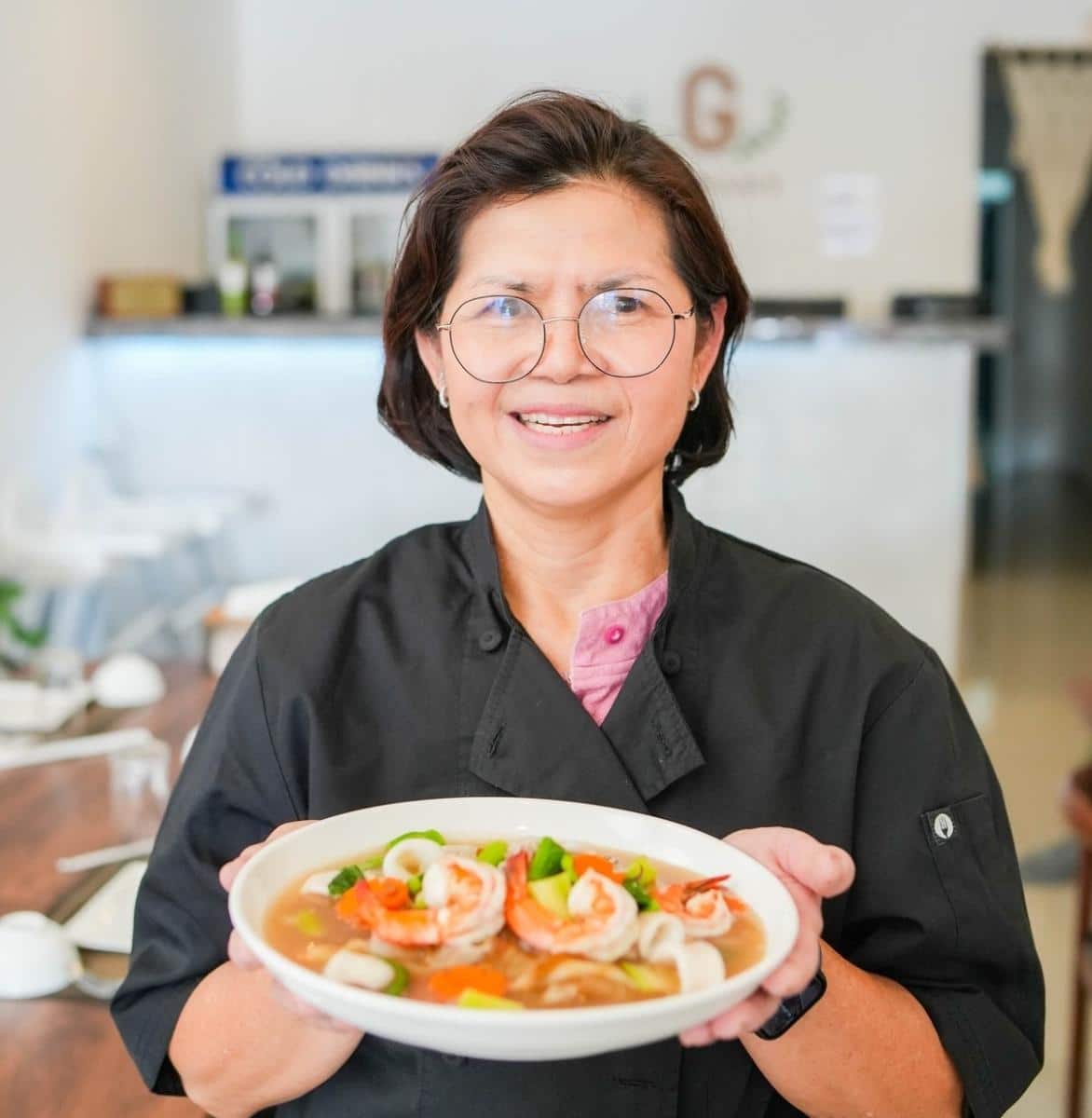 A woman in a black chef coat holds a plate of shrimp soup with vegetables, standing in a brightly lit restaurant.