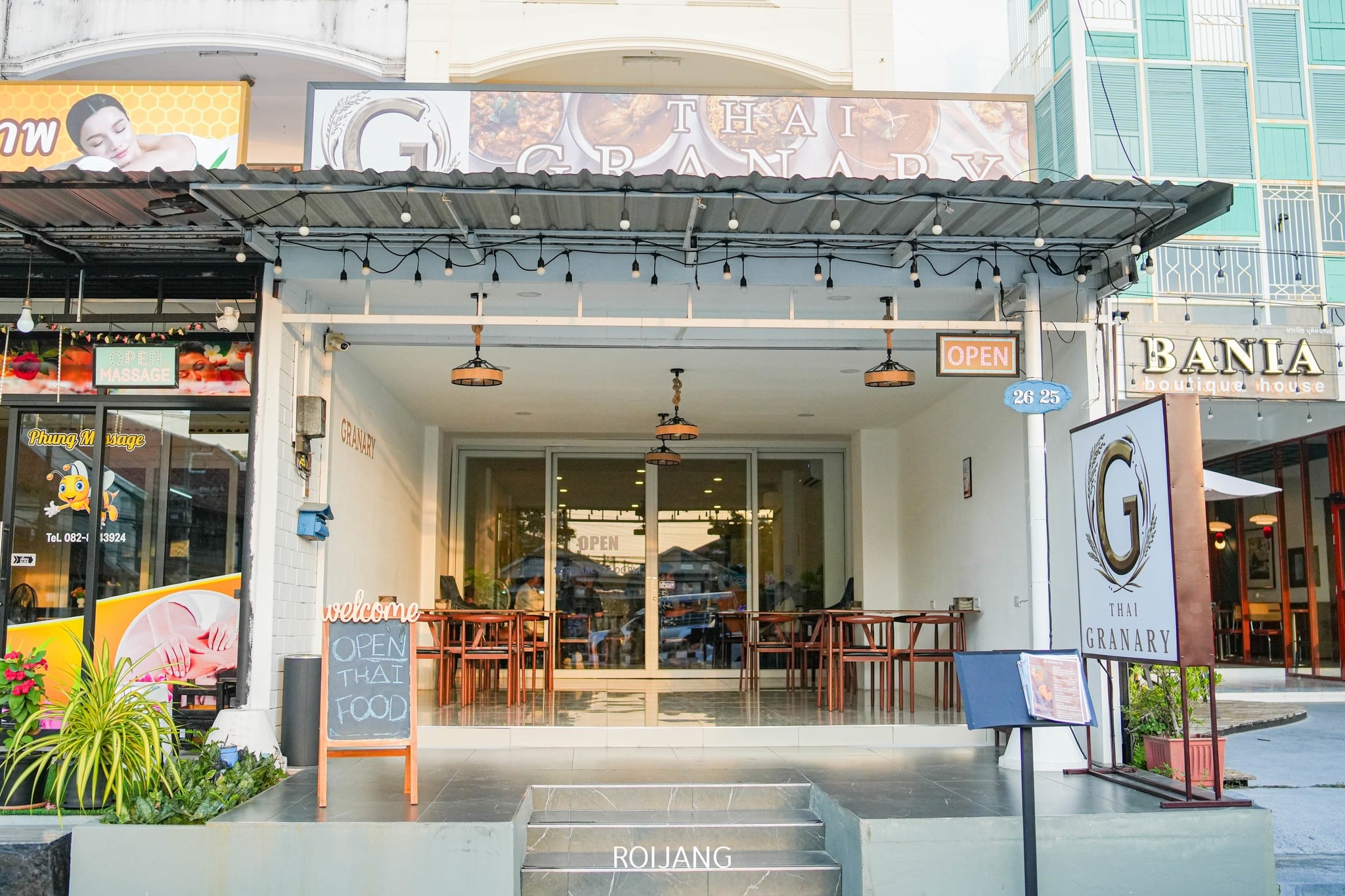 Exterior of Thai Granary restaurant with open sign, outdoor seating, and dining tables visible inside, located between other businesses on a city street.