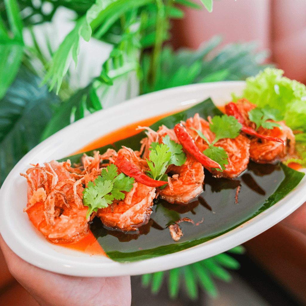 A white oval plate with five pieces of shrimp in red sauce, garnished with cilantro, fried onions, and dried chili, served on a banana leaf with lettuce on the side.