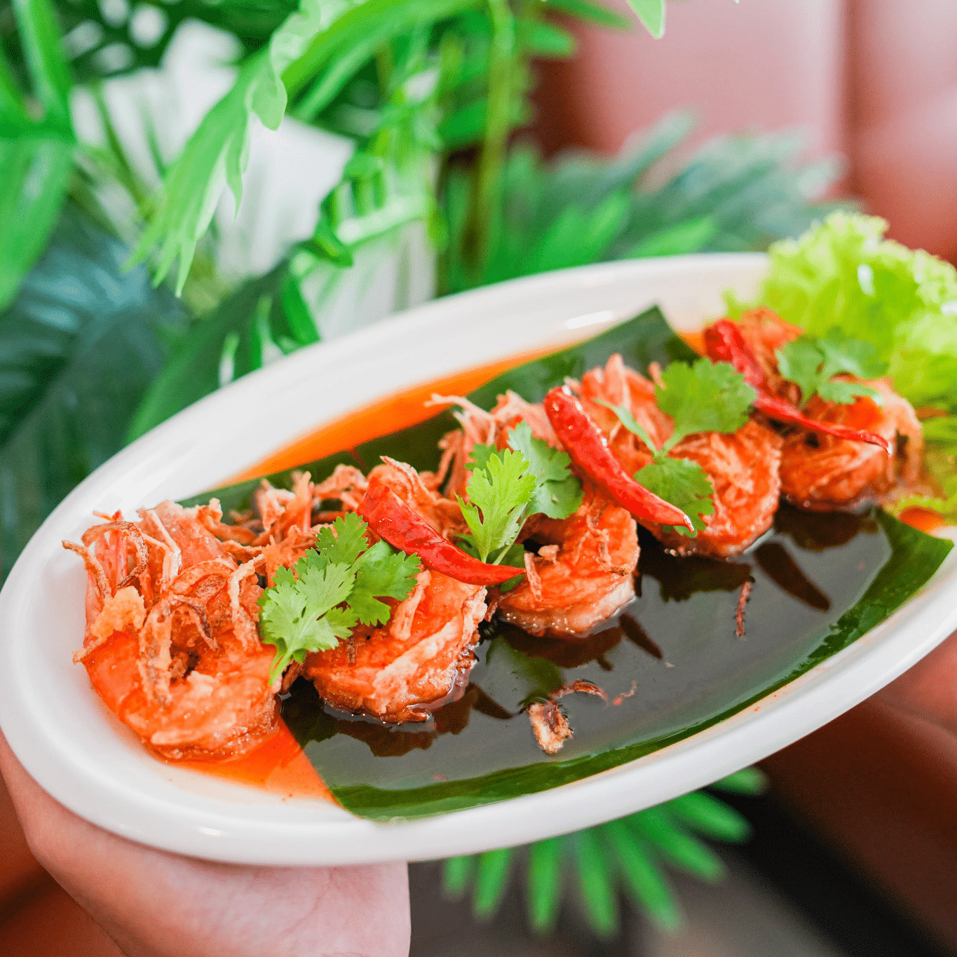 A white oval plate with five pieces of shrimp in red sauce, garnished with cilantro, fried onions, and dried chili, served on a banana leaf with lettuce on the side.
