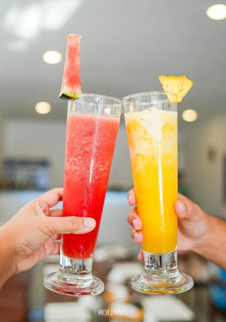 Two hands holding tall glasses of fruit drinks; one red with a watermelon garnish, the other yellow with a pineapple garnish, raised in a toast.