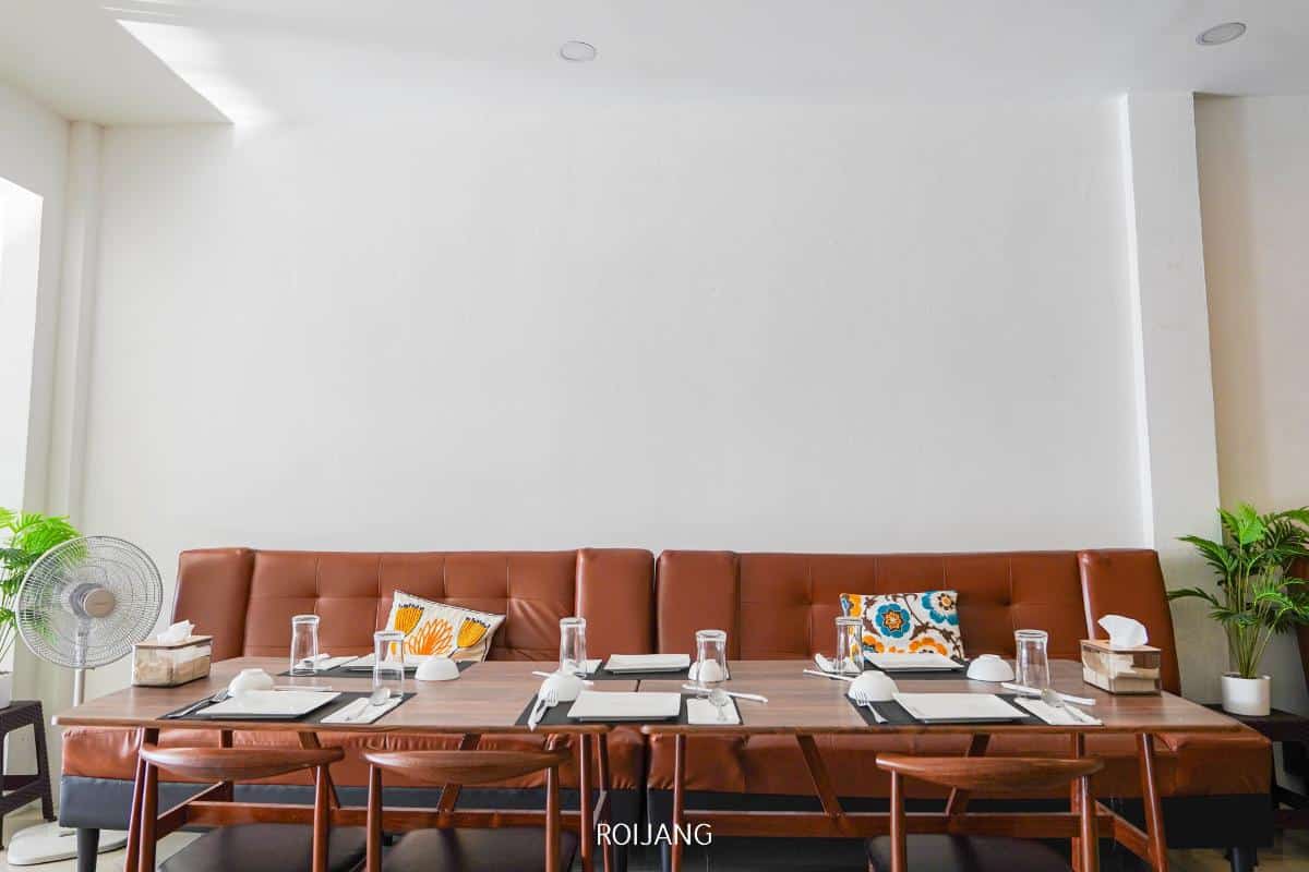 A dining table set for six with brown chairs, glassware, plates, and decorative pillows on a brown sofa against a white wall; potted plants and a fan are nearby.