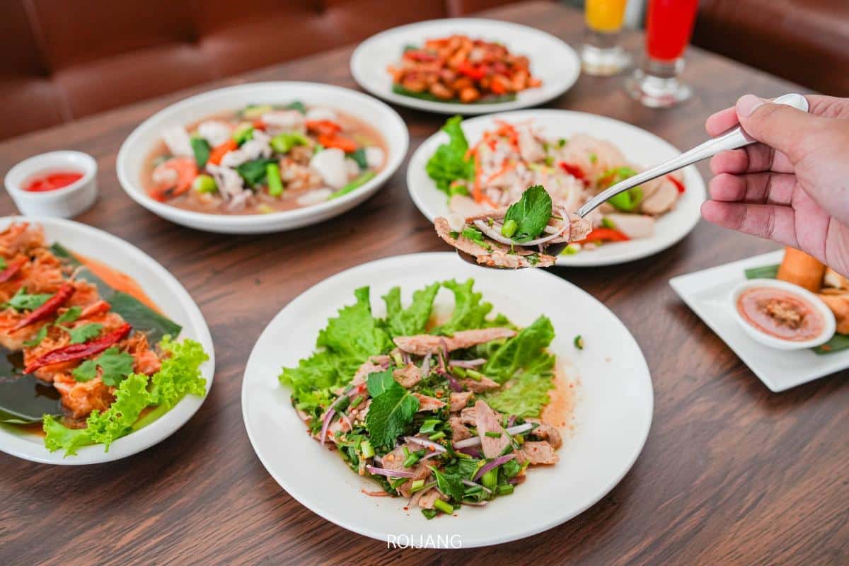 A hand holds a fork with a bite of salad over a table set with various Thai dishes, including grilled meat, salad, and vegetables.
