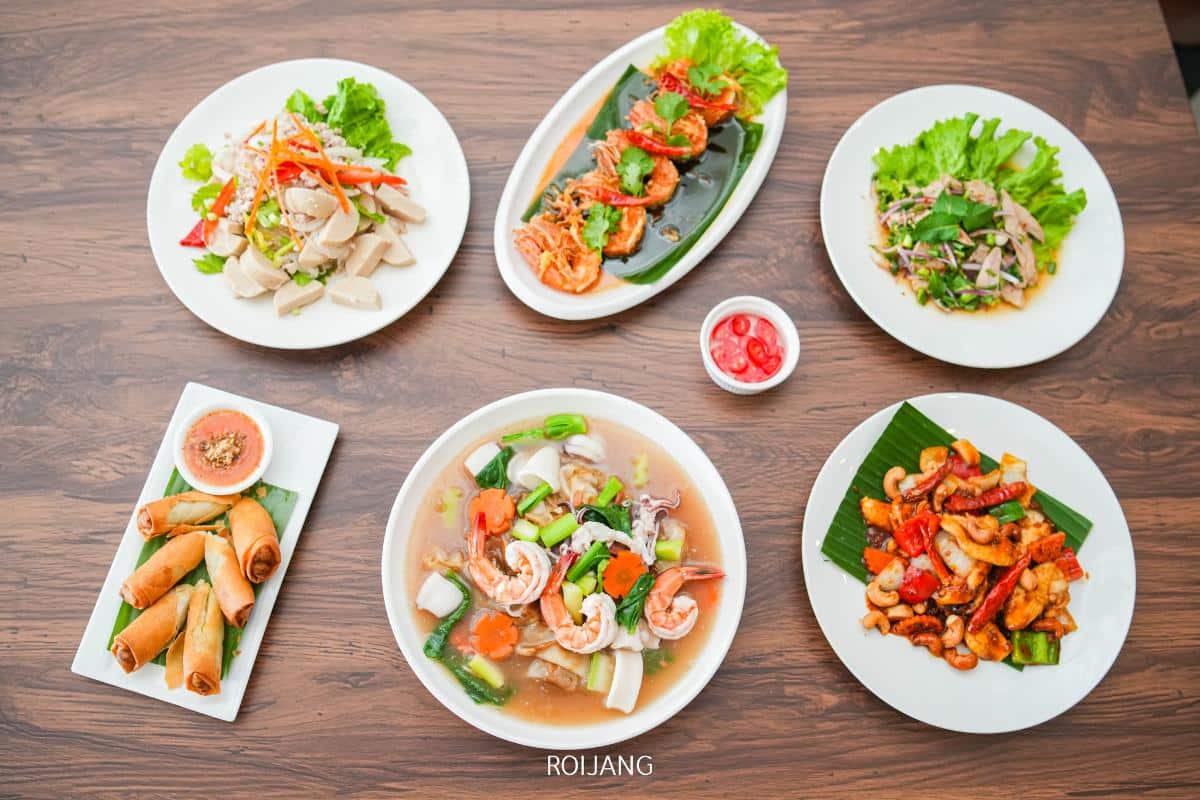 Six plates of assorted Thai dishes, including spring rolls, stir-fried shrimp, mixed vegetables with seafood, and a small bowl of cherry tomatoes, arranged on a wooden table.