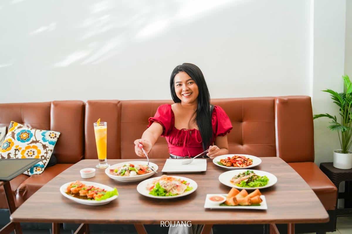 A woman sits at a table with various dishes and a drink in front of her, smiling and holding a fork, in a well-lit restaurant setting.