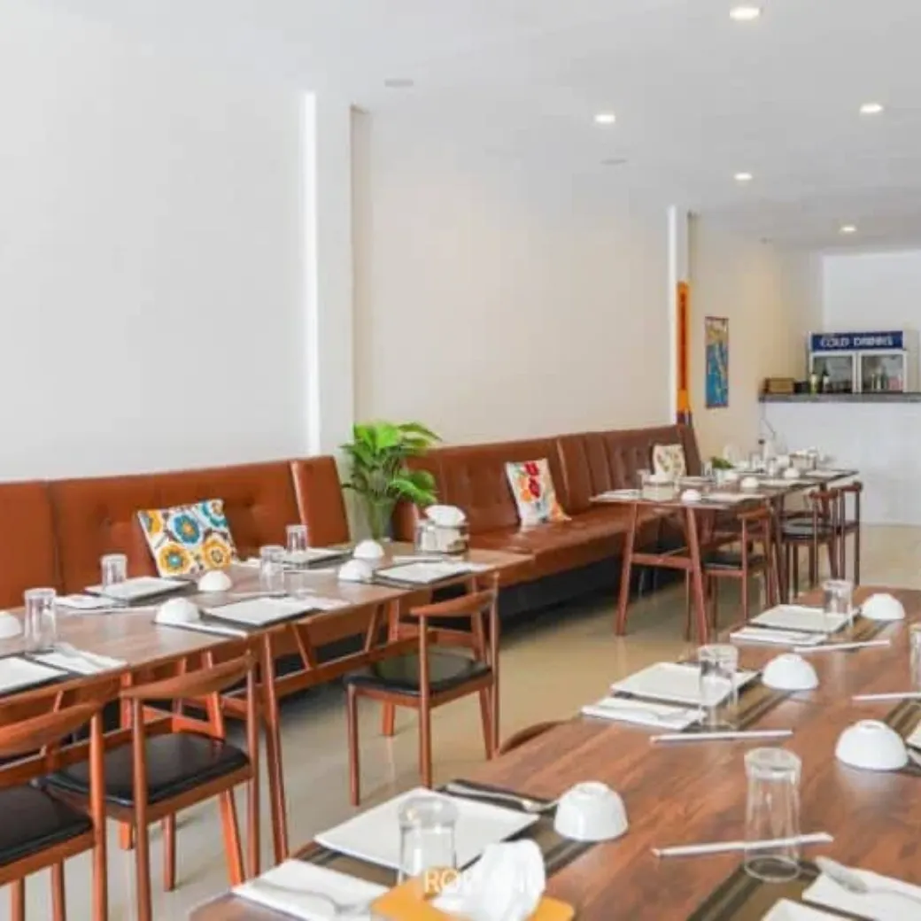 A modern restaurant interior with wooden tables set with plates, bowls, glasses, and utensils, brown bench seating, decorative cushions, and a visible counter labeled "Cold Drinks.