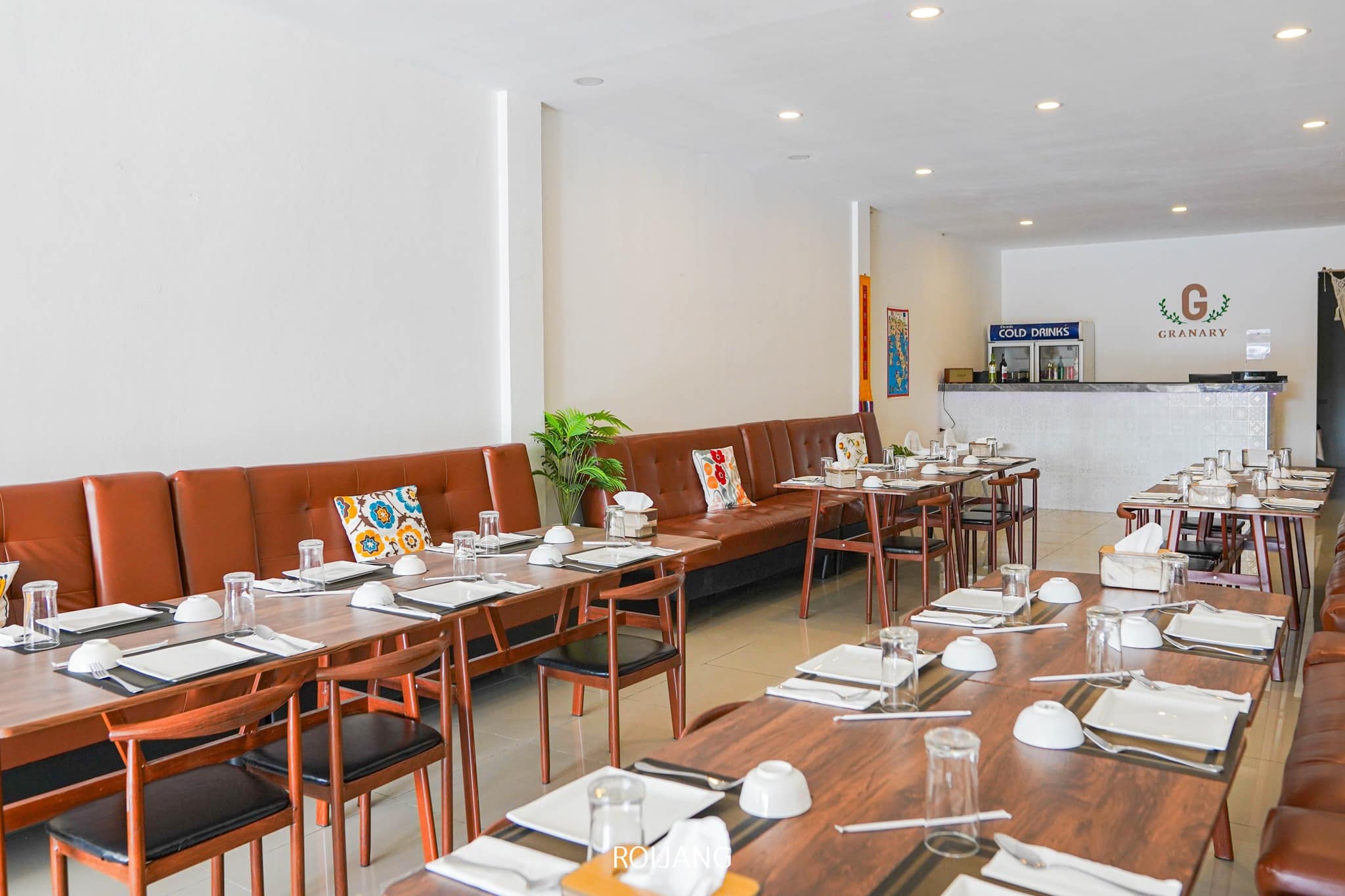 A modern restaurant dining area with wooden tables, brown cushioned seating, colorful pillows, and place settings; a counter with a "Granary" sign is visible in the background.