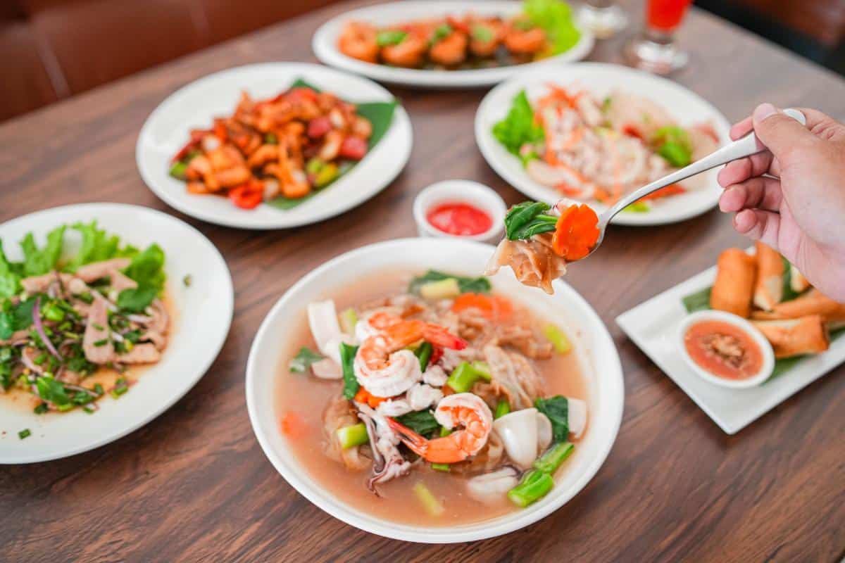 A hand holds a fork with food above a bowl of noodle soup, surrounded by various Thai dishes on a wooden table.