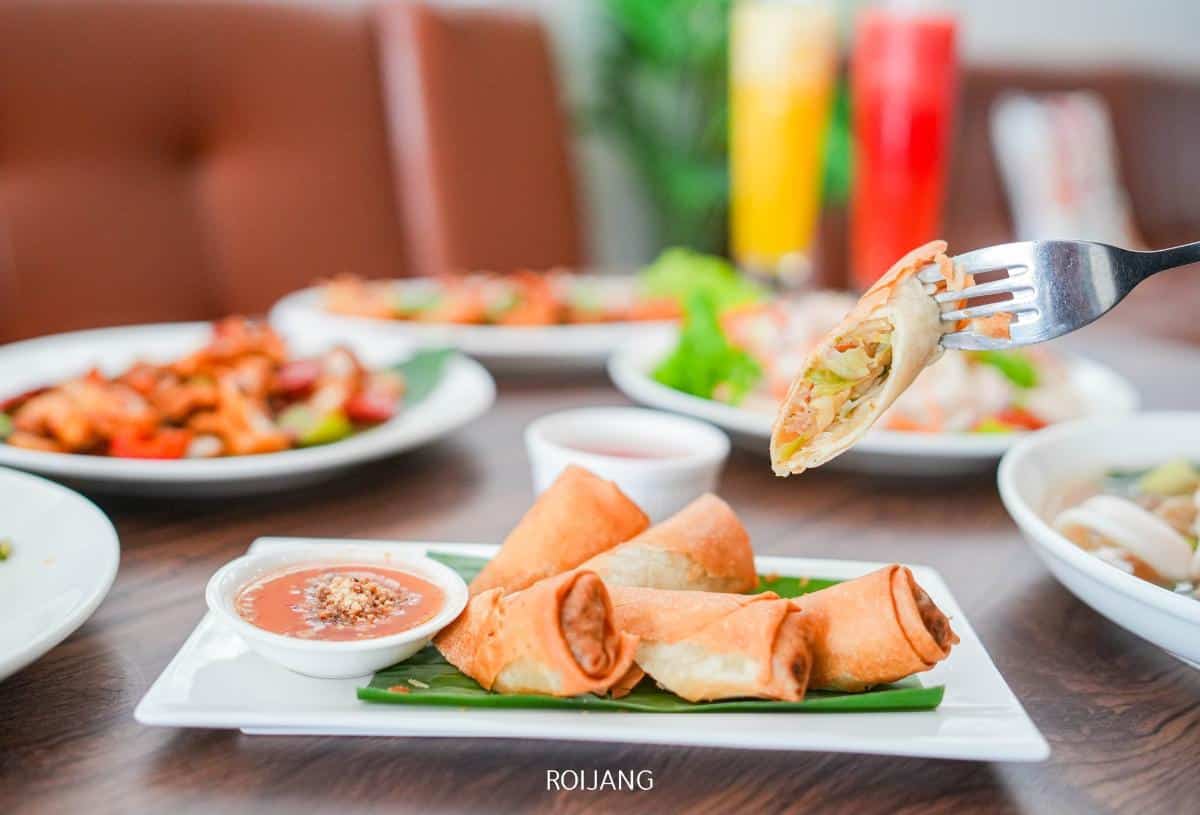 A close-up of spring rolls on a plate with dipping sauce, with one spring roll held up by a fork. Other dishes and drinks are visible in the background.