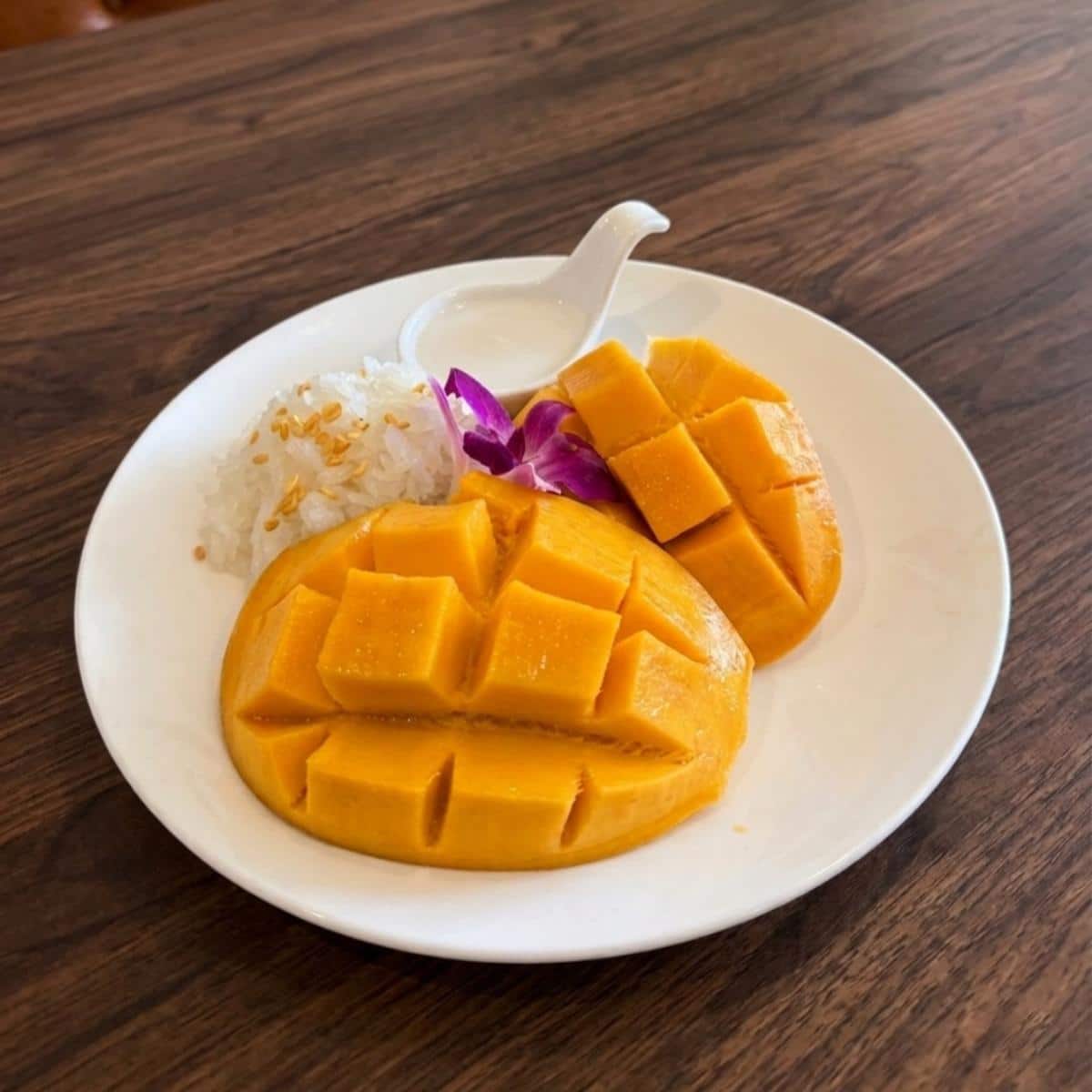 A plate with sliced mango, sticky rice topped with sesame seeds, a small container of coconut milk, and a purple flower garnish on a wooden table.
