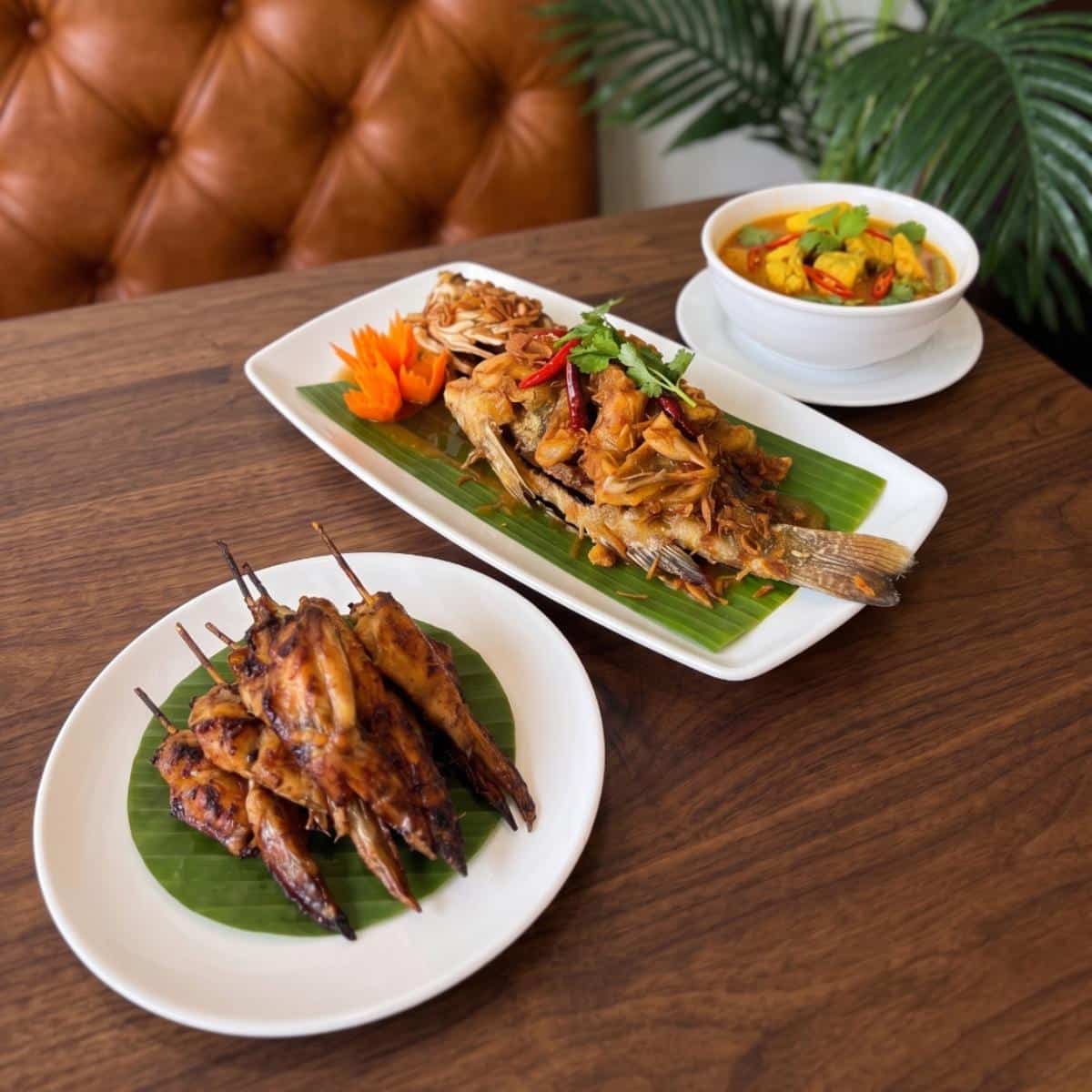 Grilled chicken skewers, a whole cooked fish with garnish, and a bowl of curry are served on a wooden table with a plant in the background.