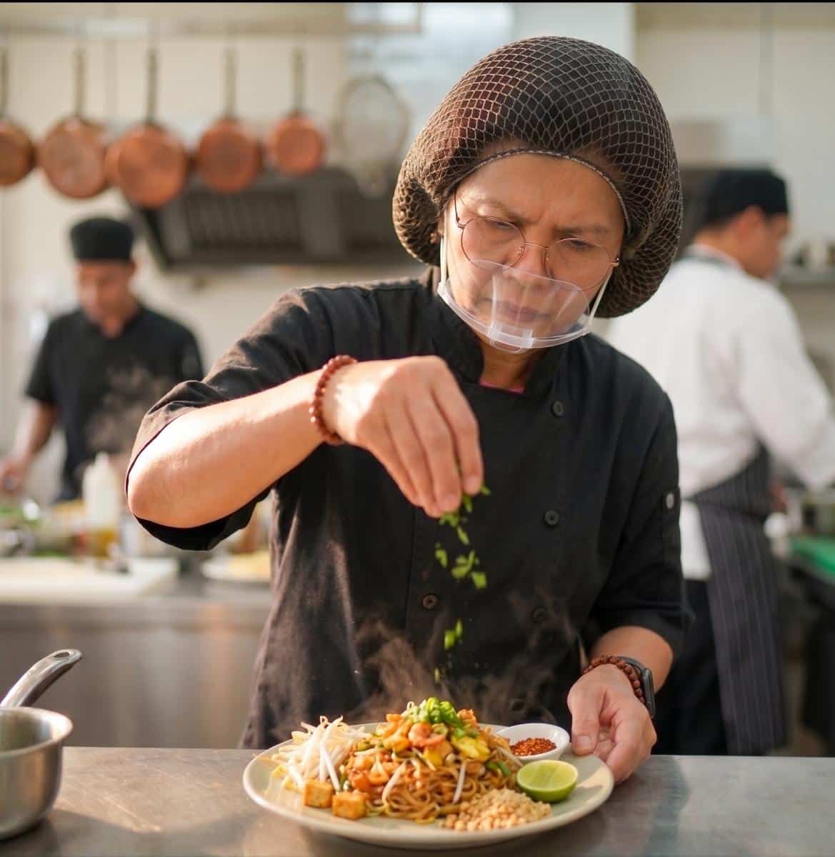 A chef wearing a hairnet and face shield sprinkles herbs onto a plate of noodles in a commercial kitchen, with other chefs working in the background.