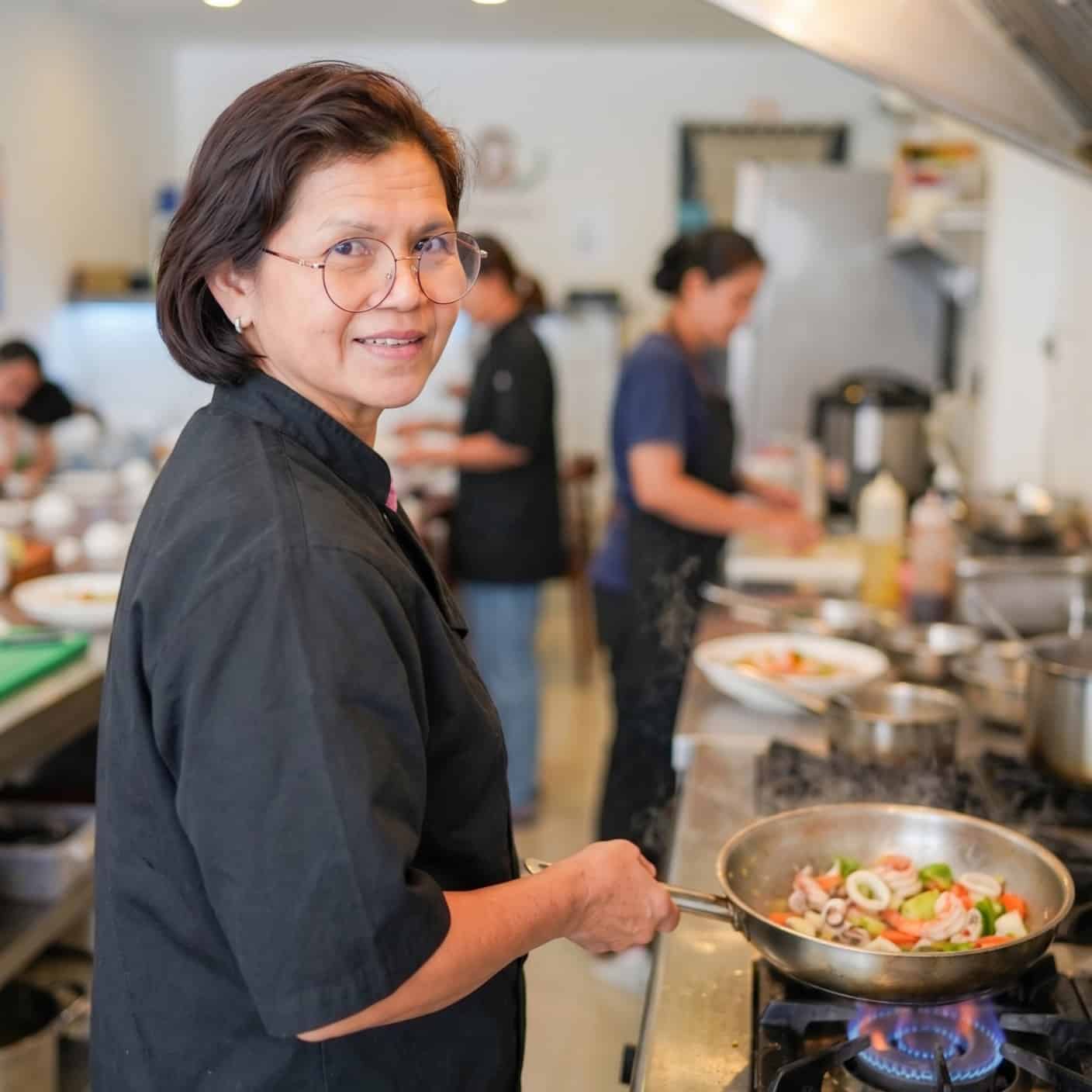 A woman in a black chef's coat cooks a stir-fry dish in a pan at a commercial kitchen, with other staff preparing food in the background.