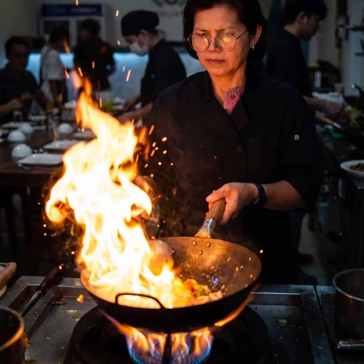 A chef stir-fries vegetables in a wok over a large open flame in a busy kitchen, with other staff and diners visible in the background.