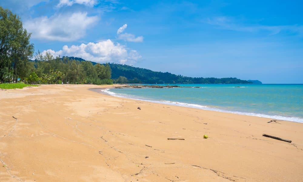 A wide, empty sandy beach with gentle waves, clear blue water, scattered driftwood, and trees along the shore under a partly cloudy sky.