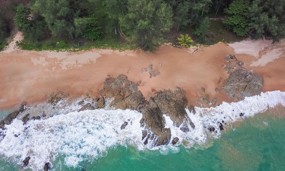 Aerial view of a sandy beach with rocky formations, waves crashing on the shore, and dense green trees lining the edge of the sand.