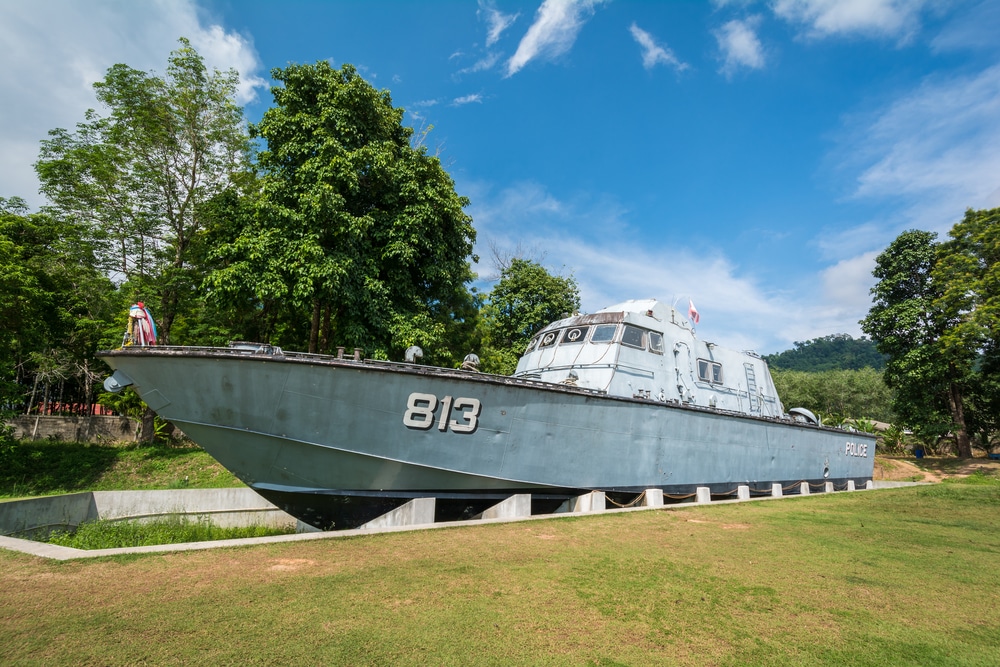 A gray police patrol boat numbered 813 is displayed on land, surrounded by grass and trees under a blue sky.